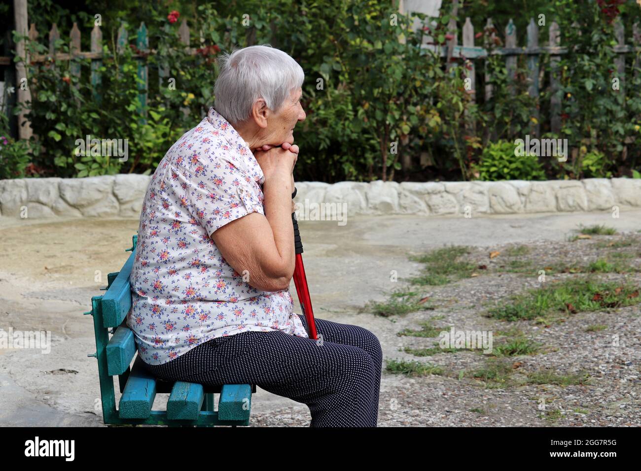 Ältere Frau, die mit Gehstock auf einer Bank in einem Hof sitzt. Gesunde Lebensweise im Alter, Leben im Ruhestand Stockfoto