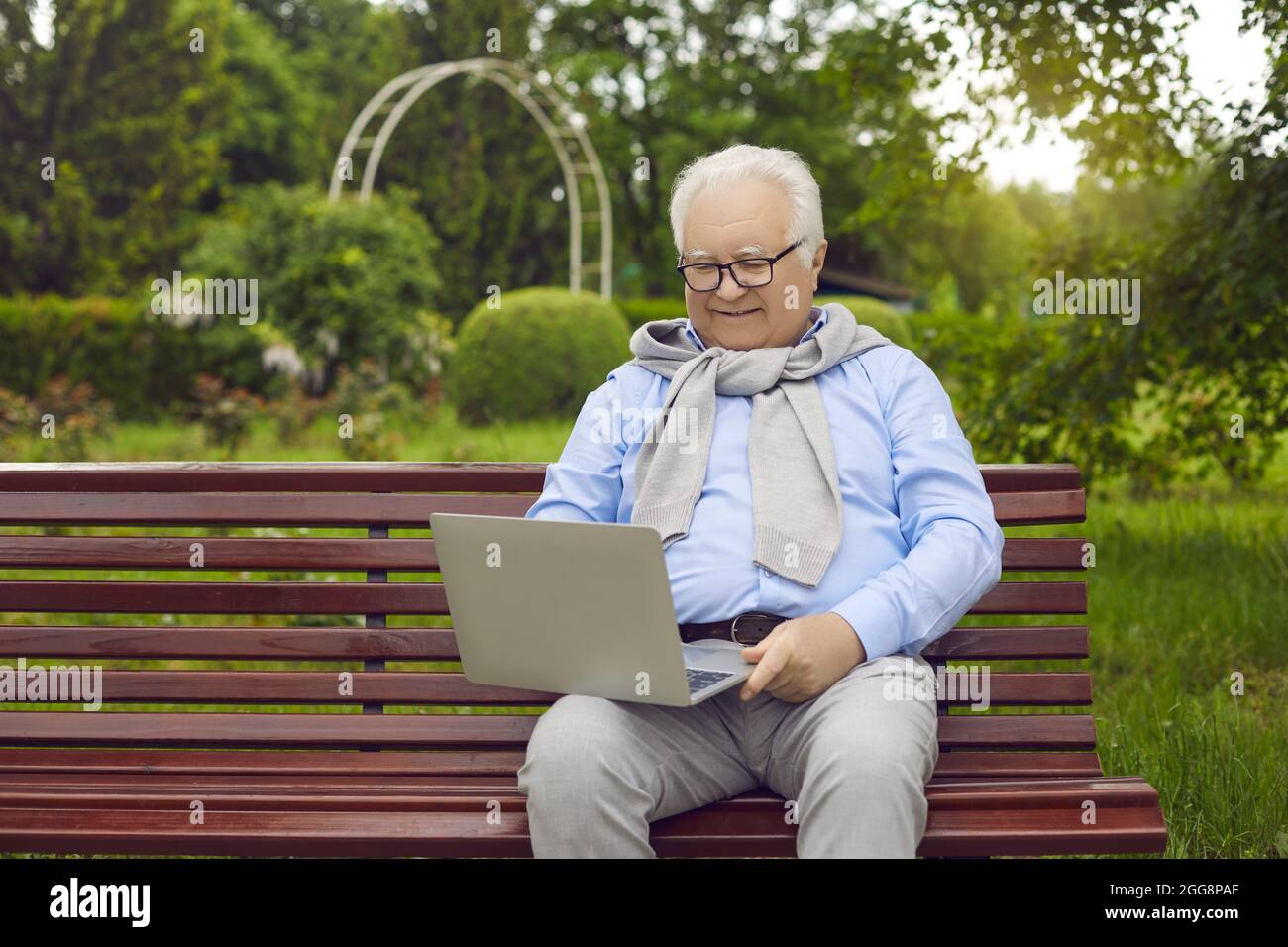 Ein älterer glücklicher Mann mit asiatischem Aussehen sitzt auf einer Bank in einem stolzen Park mit einem Laptop. Stockfoto