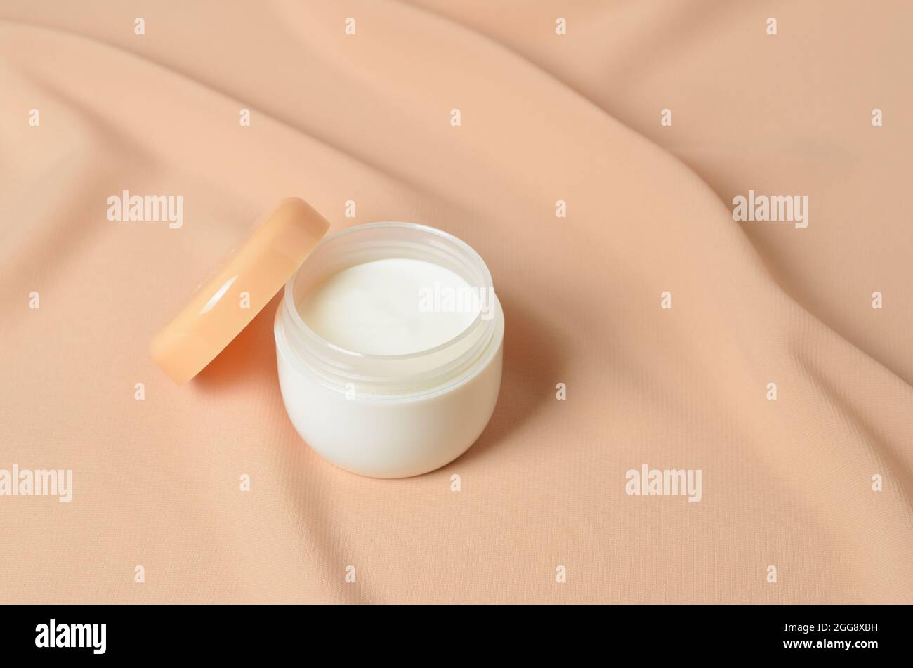 Ein Glas Creme für die Pflege zarter Haut auf einem welligen beigen Hintergrund aus Chiffon. Selektiver Fokus. Stockfoto