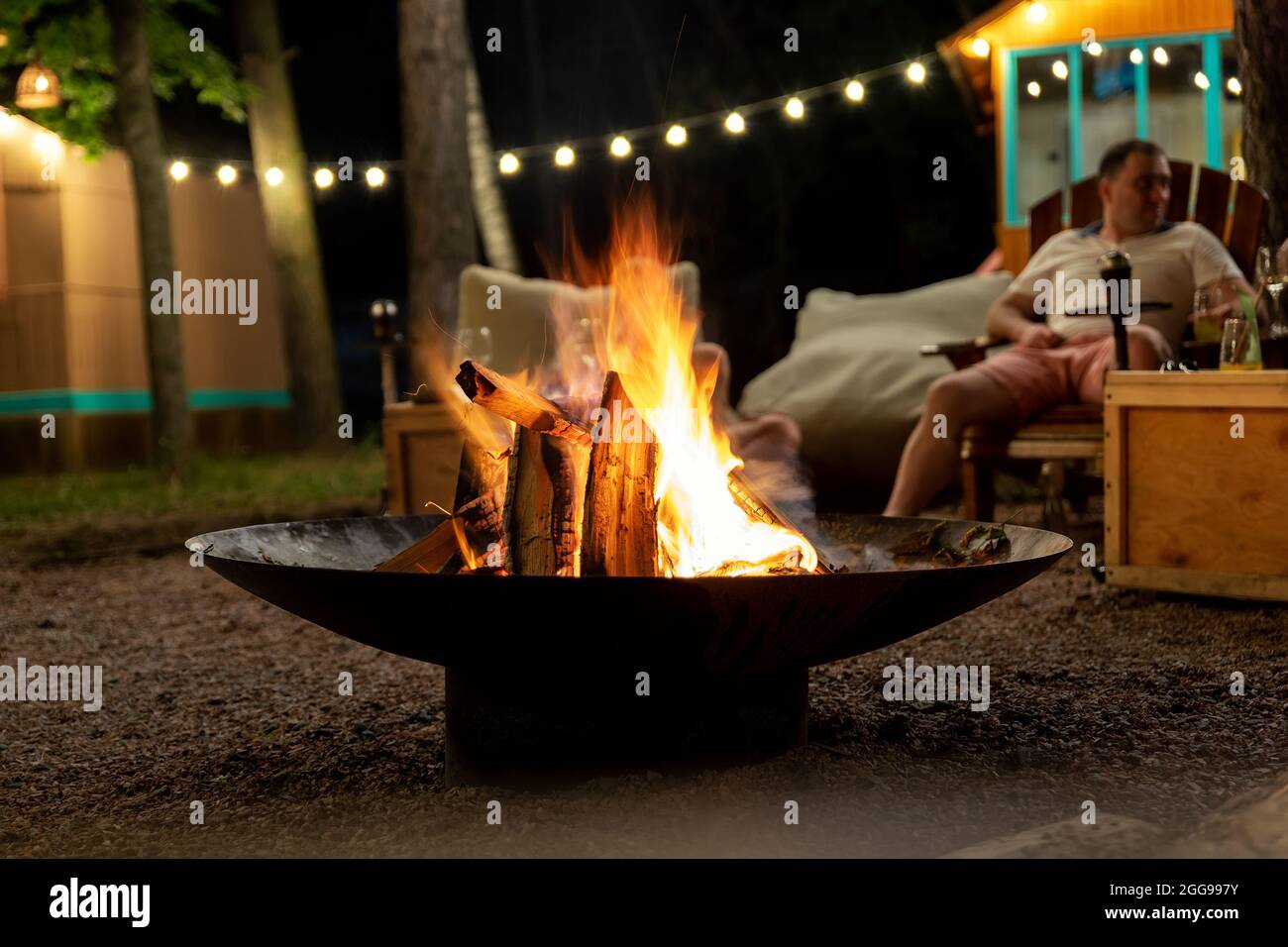 Lagerfeuer aus Gusseisen am Waldstrand Camping mit brennender Flamme am Abend gegen Glühbirnen Girlande und Bäume. Freunde sitzen Stockfoto