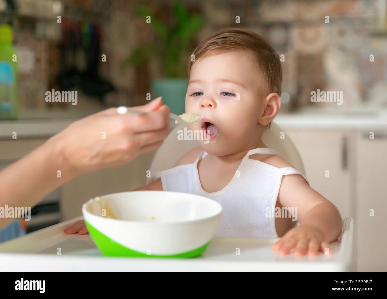 Kind füttern. Niedliches Baby, das Essen isst. Seine Mutter füttert ihn mit einem Löffel. Erster Köder. Porträt eines glücklichen jungen Kindes auf einem Hochstuhl, das gefüttert wird Stockfoto