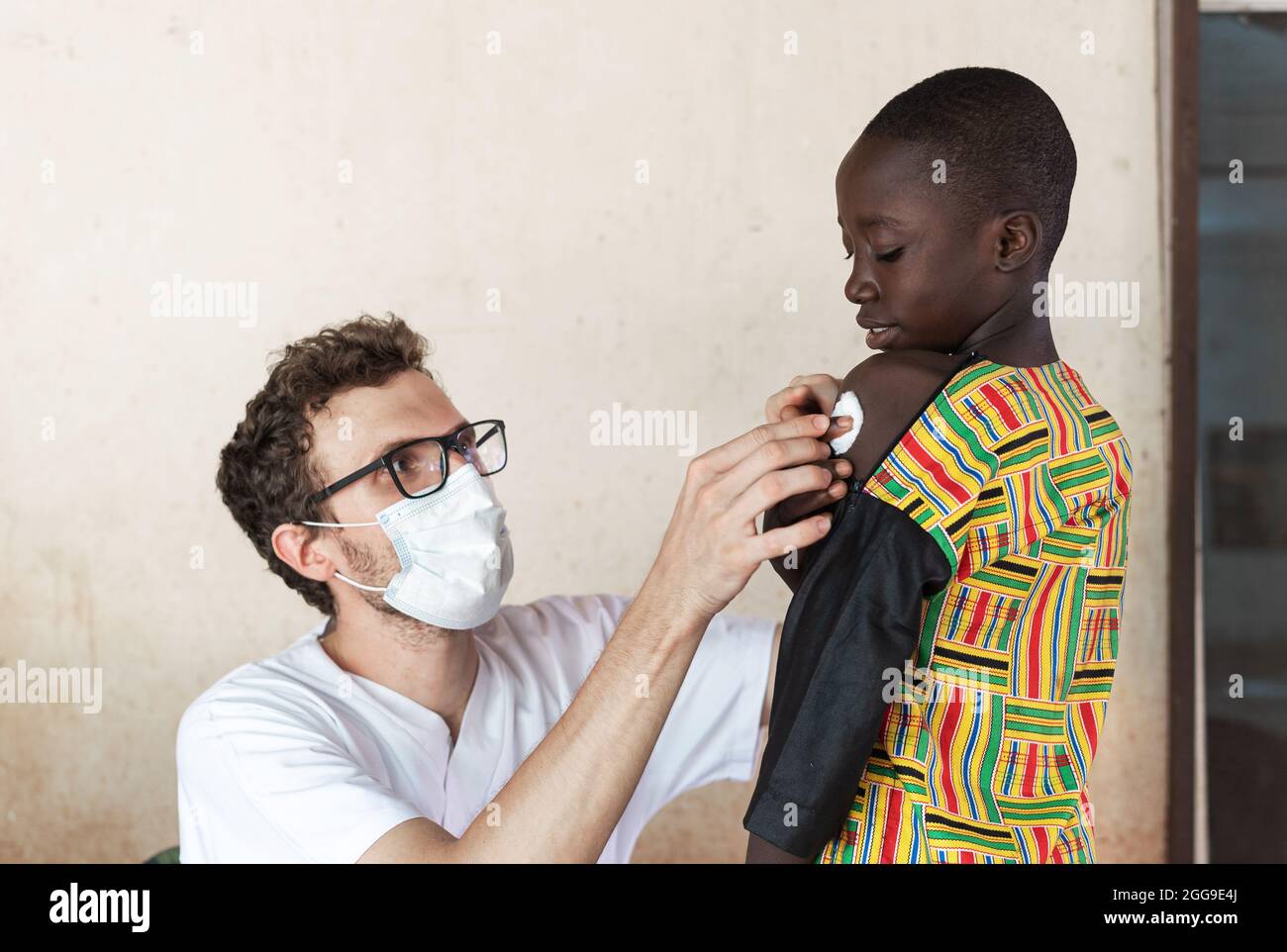 Weißer Arzt mit Gesichtsmaske und Brille, der nach der Impfung einen Wattebausch auf die Injektionsstelle am Arm eines kleinen afrikanischen Patienten legt Stockfoto