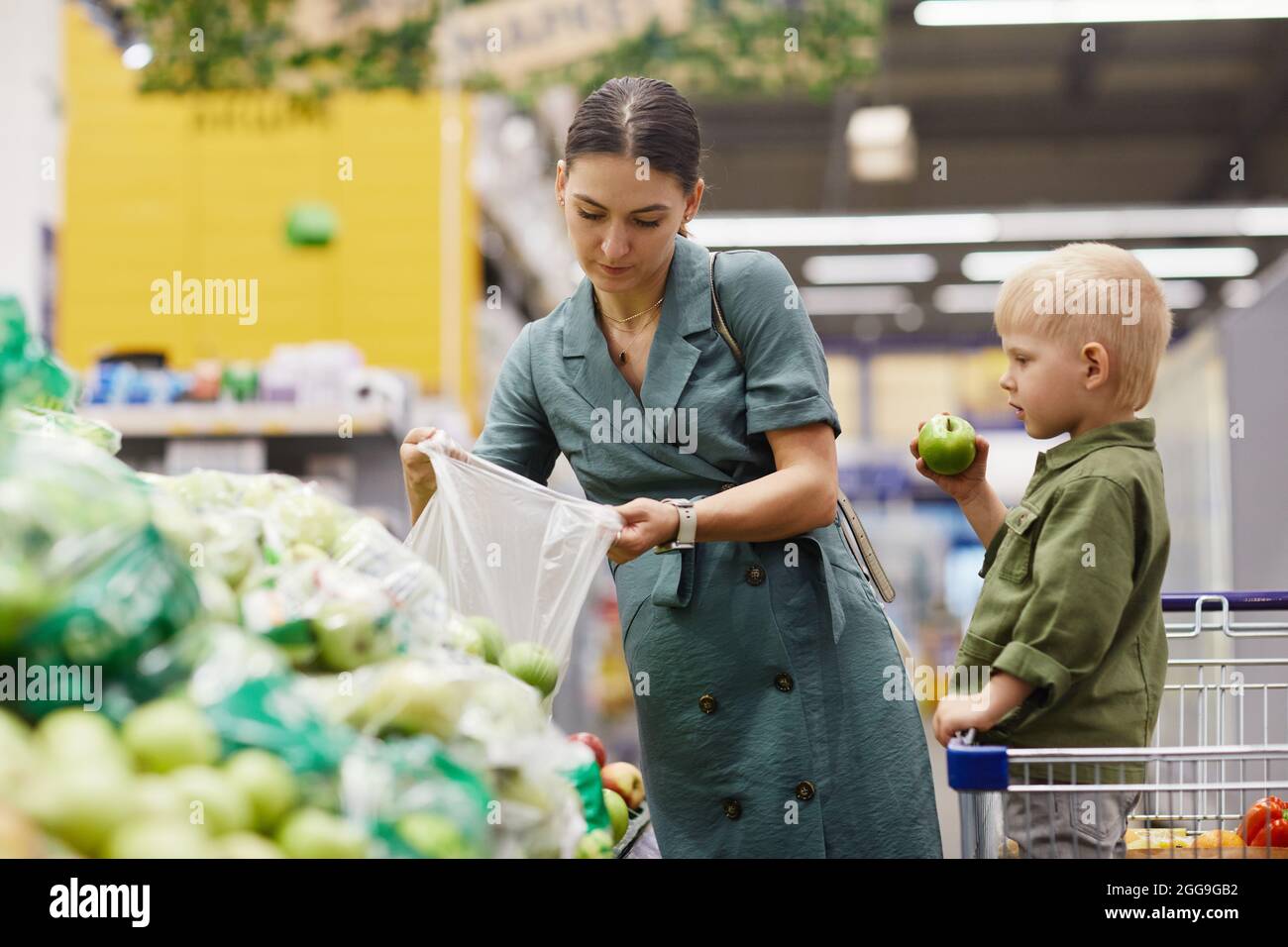 Junge Brünette Frau in Kleid setzen Äpfel in Plastiktüte, während ihr Sohn essen Apfel in den Warenkorb auf Bauernmarkt Stockfoto