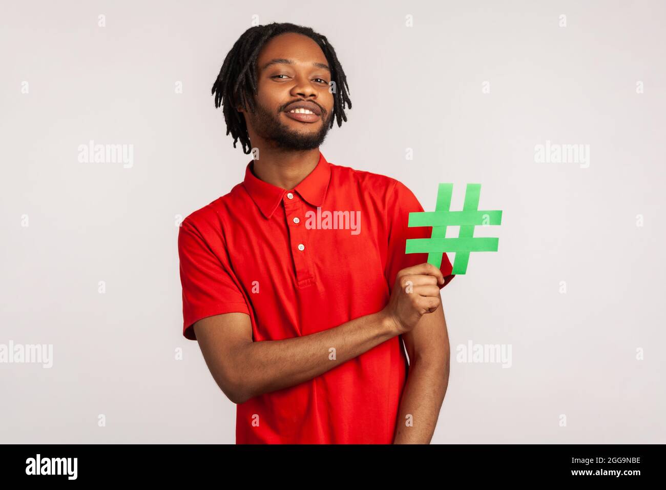 Abonnieren Sie den beliebten Blog! Bärtiger Mann mit Dreadlocks trägt ein rotes T-Shirt im lässigen Stil, hält ein Hashtag-Symbol für soziale Medien und lächelt. innenstudi Stockfoto
