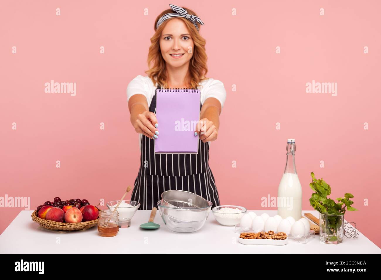 Angenehm aussehende junge Erwachsene Frau auf der Schürze, die der Kamera ein leeres Notizbuch zeigt, in der Umgebung mit Zutaten für Gebäck, Bäckerei posiert. Innen Stockfoto