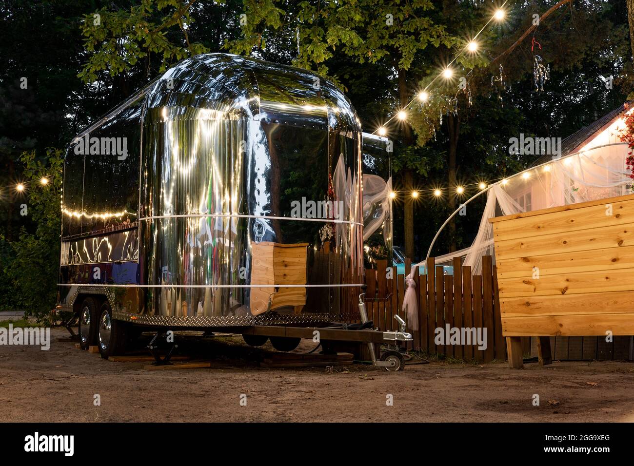 Szenische Abendansicht des futuristischen silbernen Wohnmobil-Anhängers aus Metall, der in der Nähe eines Holzzauns auf einer Farm oder Ranch vor leichter Girlande geparkt ist Stockfoto
