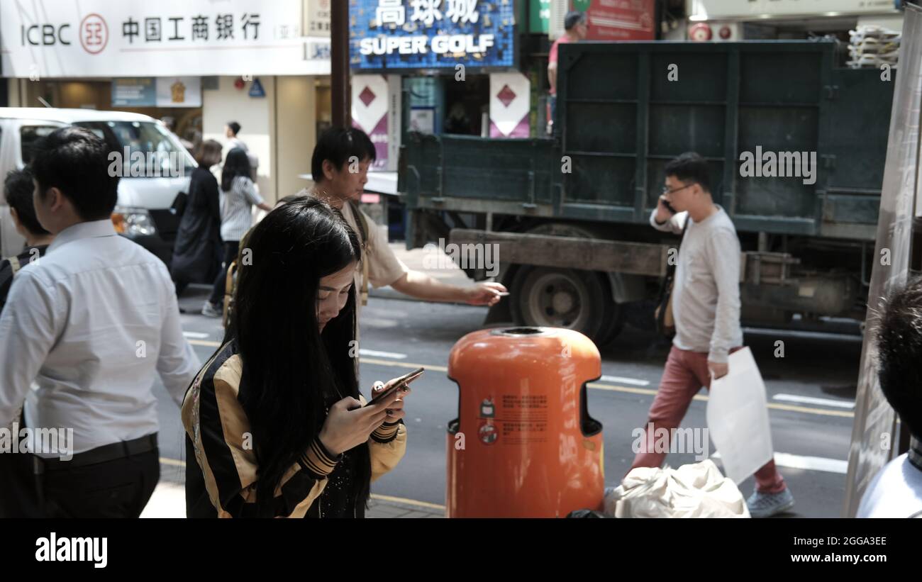 Mann mit Abfalleimer,Mädchen auf Handy, Nachbarschaft Street Scene Tsim Sha Tsui, Kowloon, Hongkong, China Stockfoto