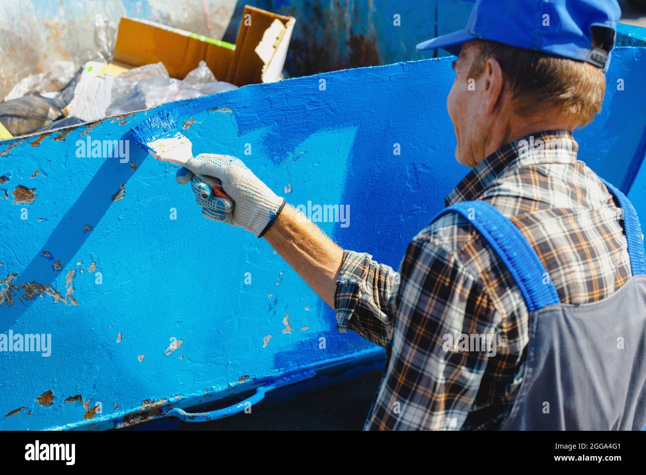 Ein alter Mann in einem Arbeitsanzug malt mit einem Pinsel auf der Straße eine Mülltonne oder einen Behälter. Teilzeitarbeit für einen Rentner. Mühsal. Stockfoto