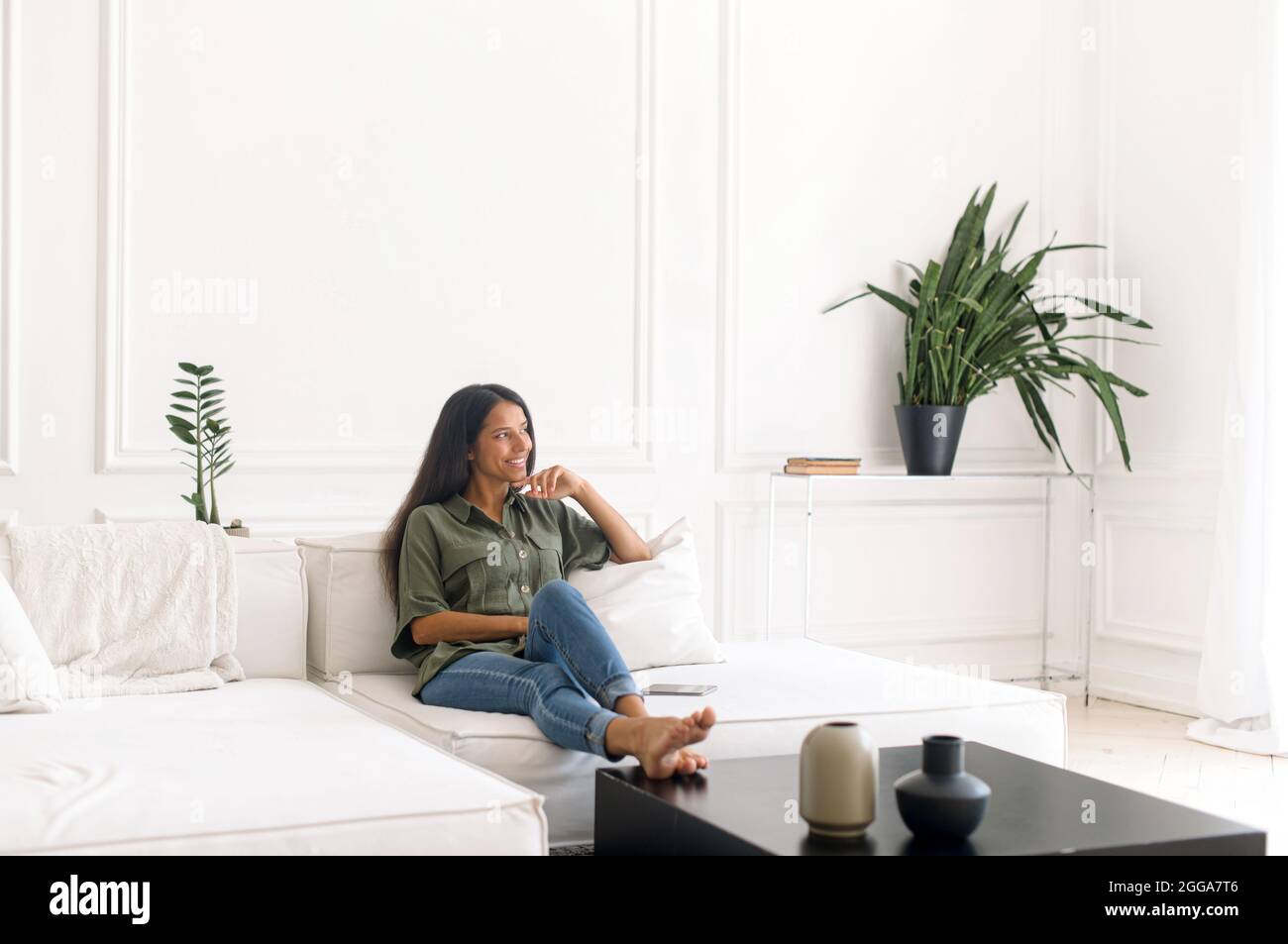 Sorglos und ruhig Attraktive junge inderin, die in entspannter Atmosphäre auf der gemütlichen weißen Couch sitzt, genießt die lächelnde Mixed-Race-Lady ein faules Wochenende zu Hause und träumt allein Stockfoto