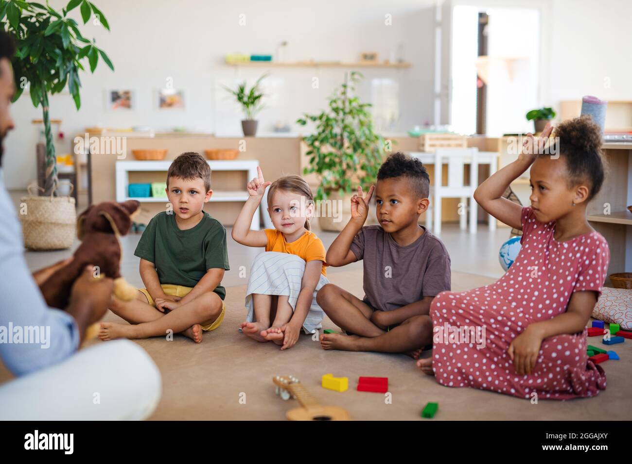 Gruppe von kleinen Kindern im Kindergarten mit einem Lehrer, der im Klassenzimmer auf dem Boden sitzt und die Hände hebt. Stockfoto