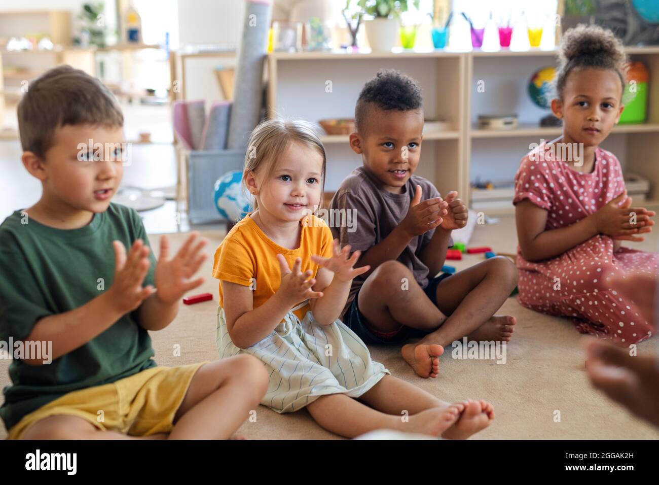 Gruppe von kleinen Kindergartenkindern, die im Klassenzimmer auf dem Boden sitzen und klatschen. Stockfoto