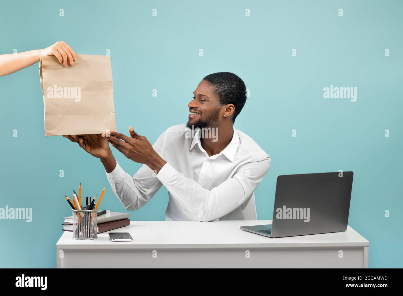 Millennial schwarzer Geschäftsmann, der die Lieferung von Lebensmitteln vom Kurier erhält, sitzt am Arbeitsplatz über blauem Studiohintergrund Stockfoto