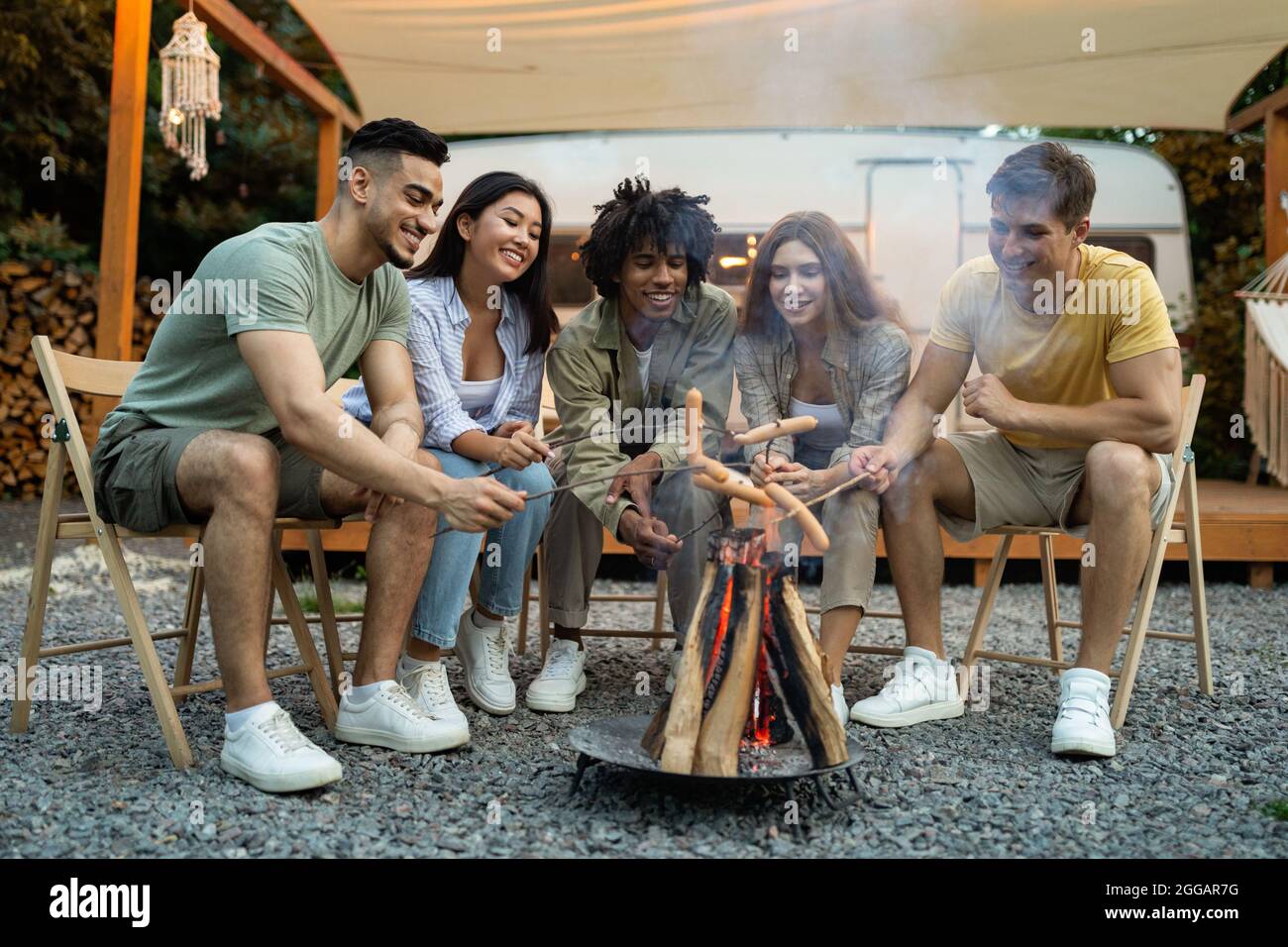 Eine Gruppe von verschiedenen Millennials, die am Lagerfeuer Würstchen braten, Picknick in der Nähe des Wohnmobils auf dem Campingausflug im Herbst Stockfoto