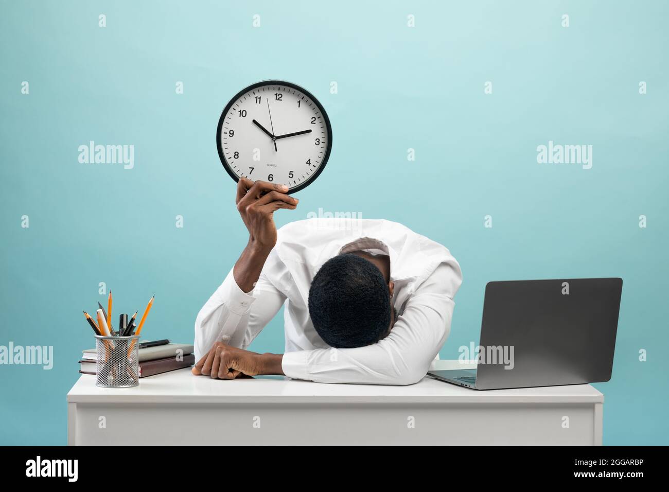 Müde afroamerikanische Geschäftsmann sitzt am Schreibtisch mit pc-Laptop, hält die Uhr, fühlt sich erschöpft, blauer Hintergrund Stockfoto
