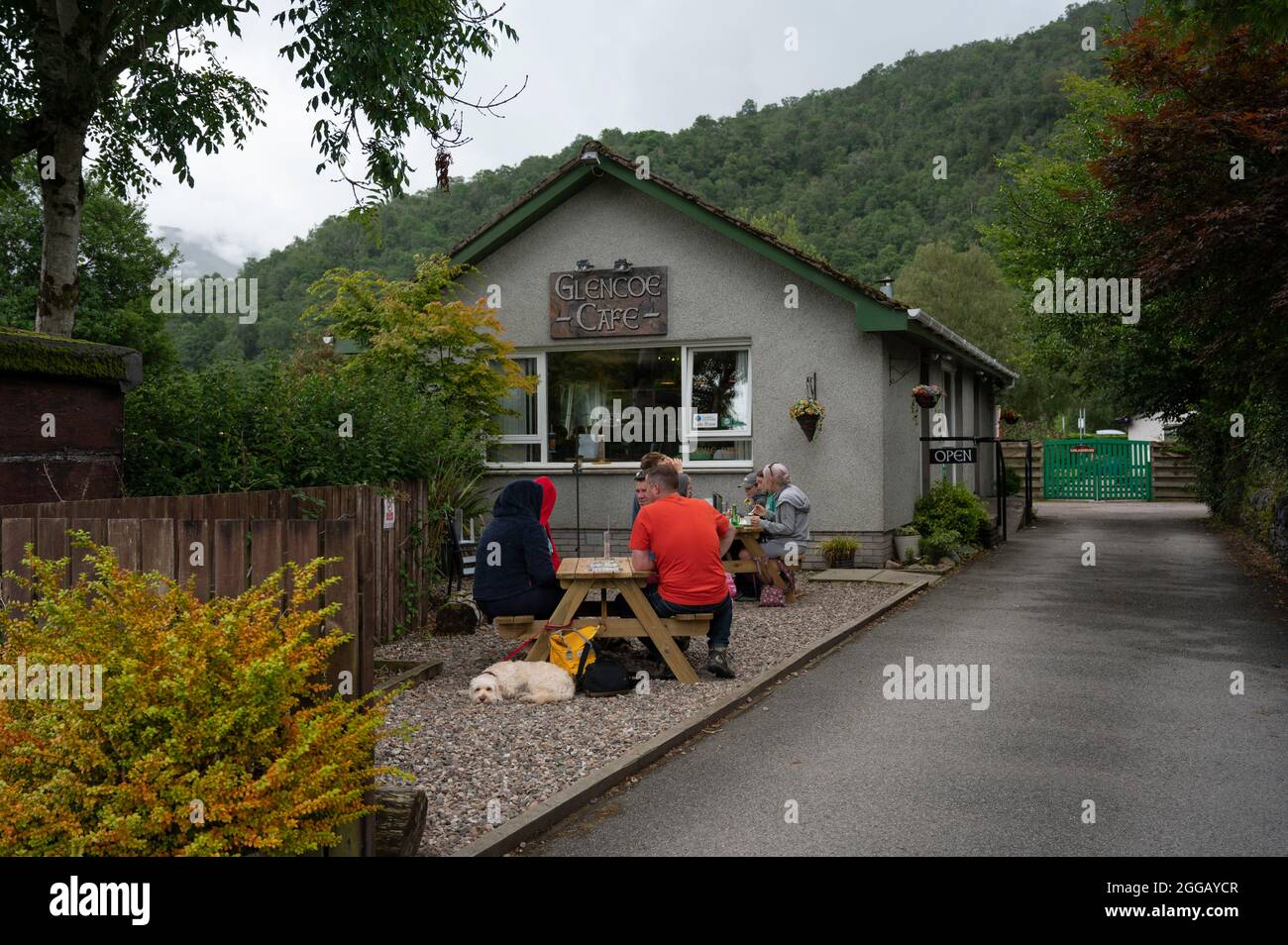 Außenansicht des Glencoe Cafe in den schottischen Highlands. Menschen und Hund essen draußen auf Bänken. Cafe-Schild und fahren zum Parkplatz. Stockfoto