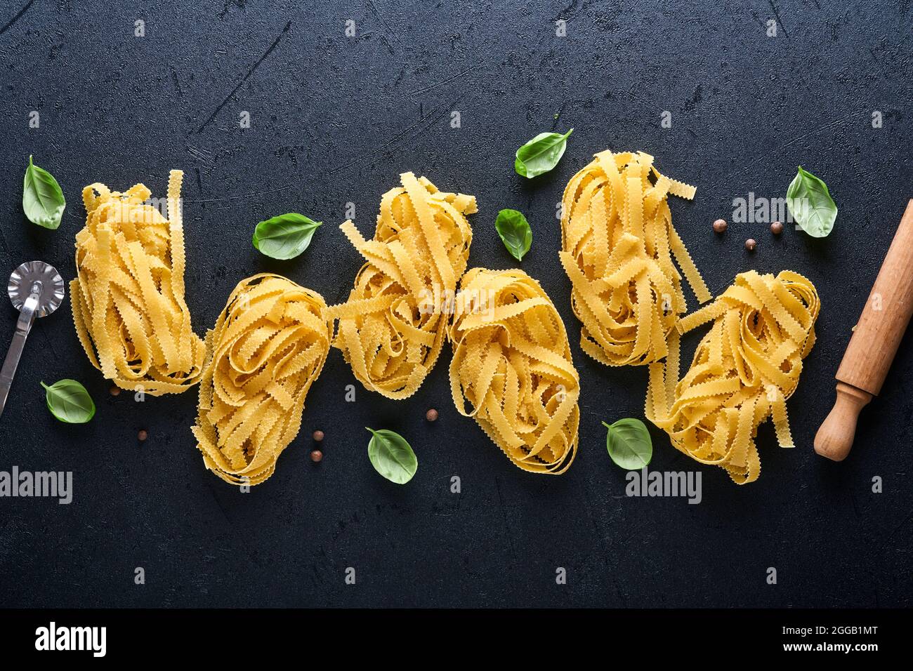 Tagliatelle. Hausgemachte Pasta, Basilikumblätter, Mehl, Pfeffer, Olivenöl und Kirschtomate auf dunkelschwarzem Hintergrund. Lebensmittelkonzept. Modell. Horizontal mit Stockfoto