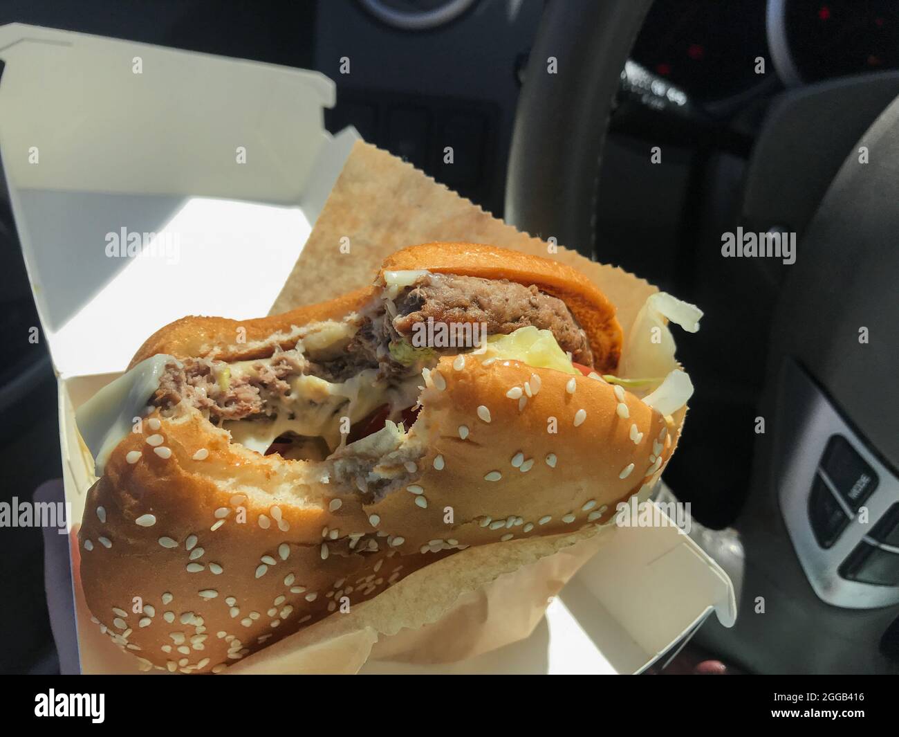 Essen während der Fahrt: Ein gebissene Burger in den Händen des Fahrers im Auto vor dem Hintergrund des Lenkrads. Stockfoto