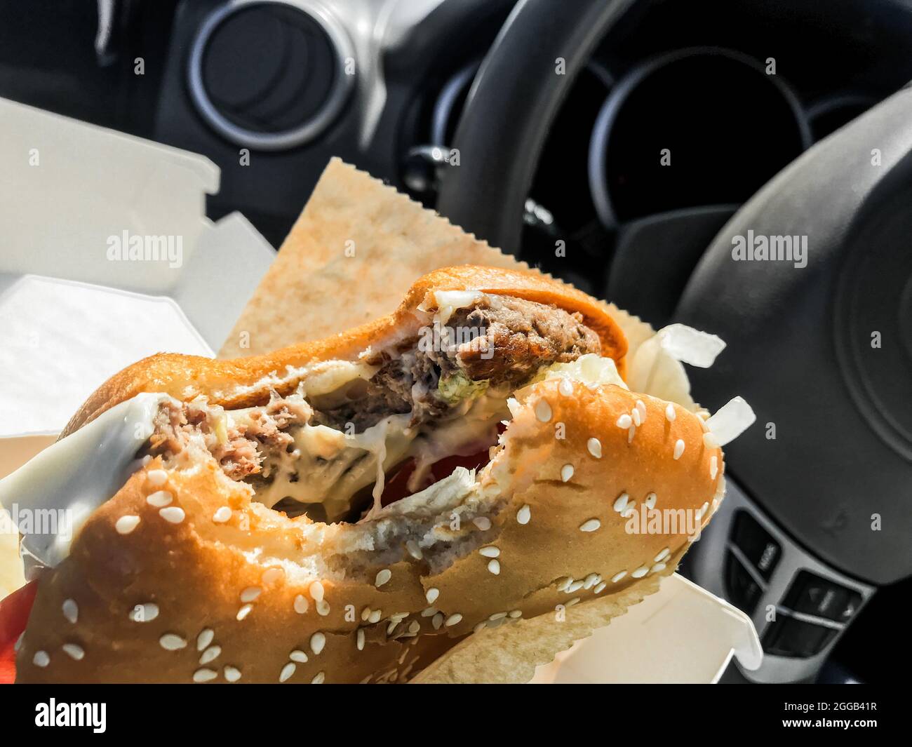 Essen während der Fahrt: Ein gebissene Burger in den Händen des Fahrers im Auto vor dem Hintergrund des Lenkrads. Stockfoto
