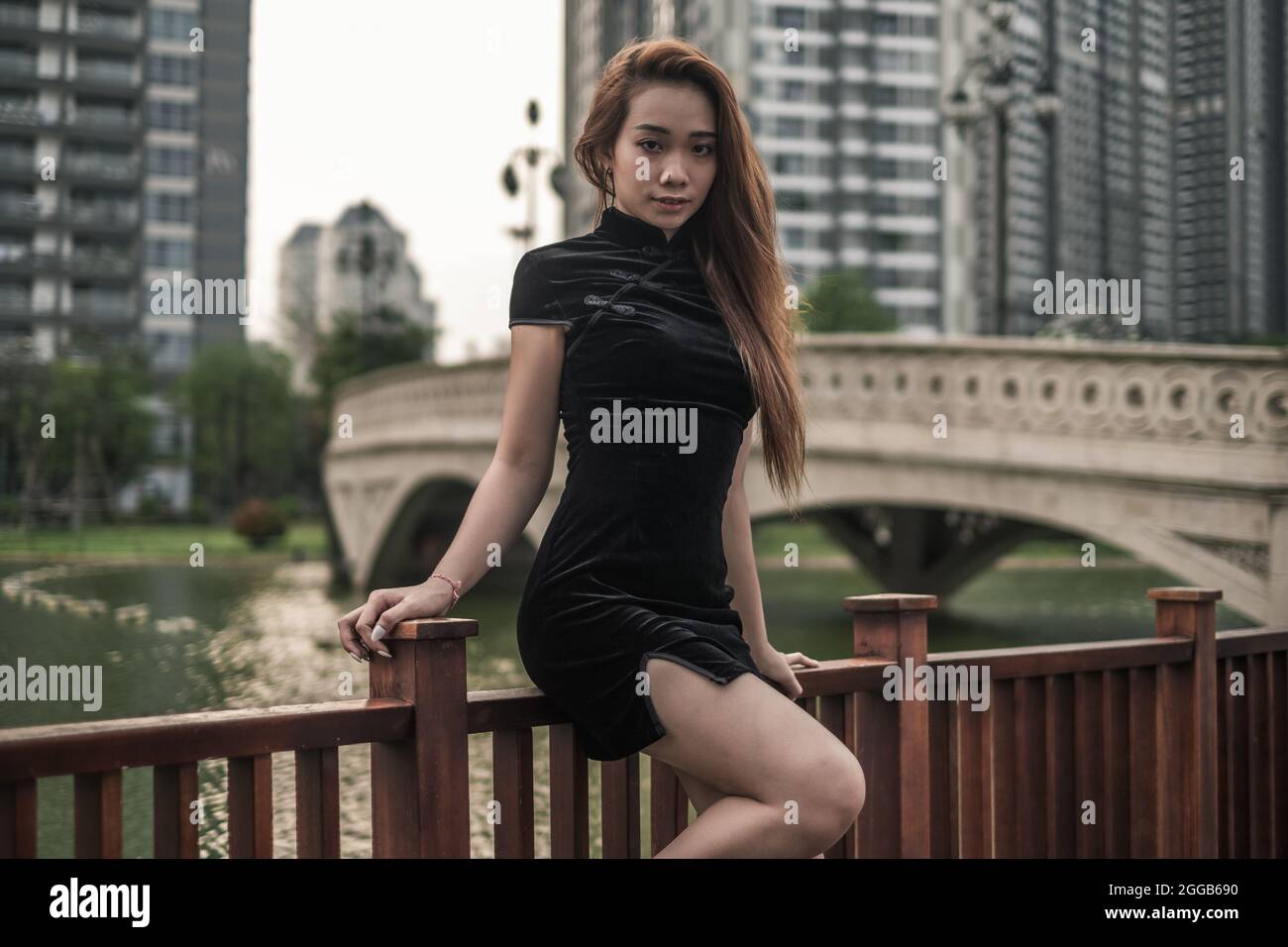 Schöne junge asiatische Frau in schwarzem Minikleid, die in der Nähe des Handlaufs im Park steht. Langes Haar. Schöne Haltung. Kamera mit Kopierplatz betrachten. Stockfoto