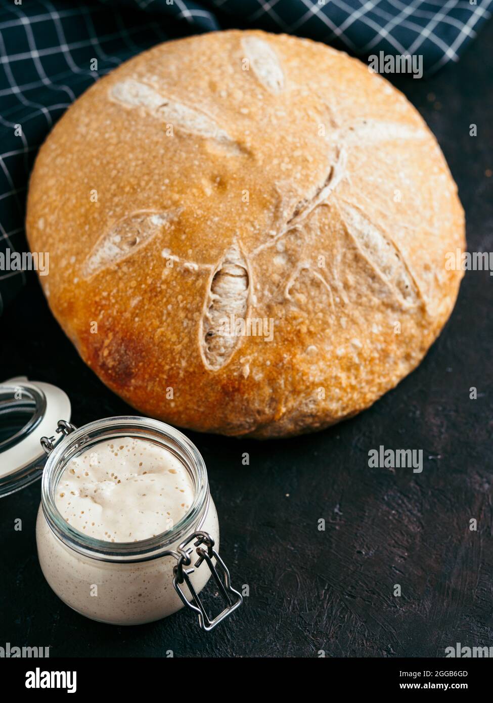 Sauerteig-Vorspeise und Weizensauerteig-Brot. Weizensaurer Teigstarter im Glasgefäß und hausgemachtes rundes Sauerteigbrot auf schwarzem Hintergrund, Kopierraum. Hausgemachter Sauerteig Brot machen Konzept Vertikal. Stockfoto