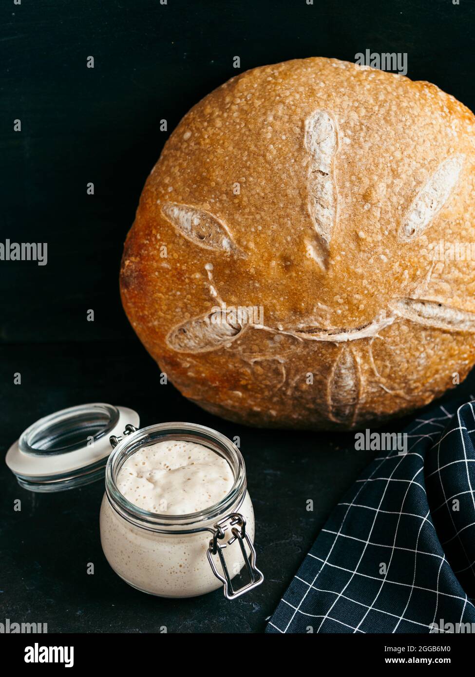 Sauerteig-Vorspeise und Weizensauerteig-Brot. Weizensaurer Teigvorspeise im Glasgefäß und leckeres hausgemachtes rundes Sauerteigbrot auf schwarzem Hintergrund. Hausgemachtes Sauerteig Brot machen Konzept. Vertikal Stockfoto