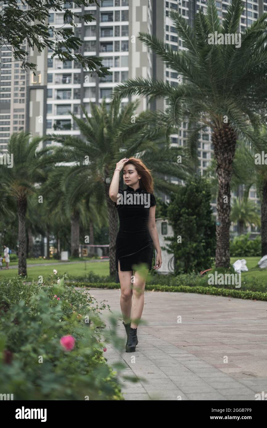 Schöne junge asiatische Frau in lässigen schwarzen Kleid zu Fuß auf der Straße mit Palmen. Langes Haar. Schöne Haltung. Reizende Vietnamesin. Stockfoto