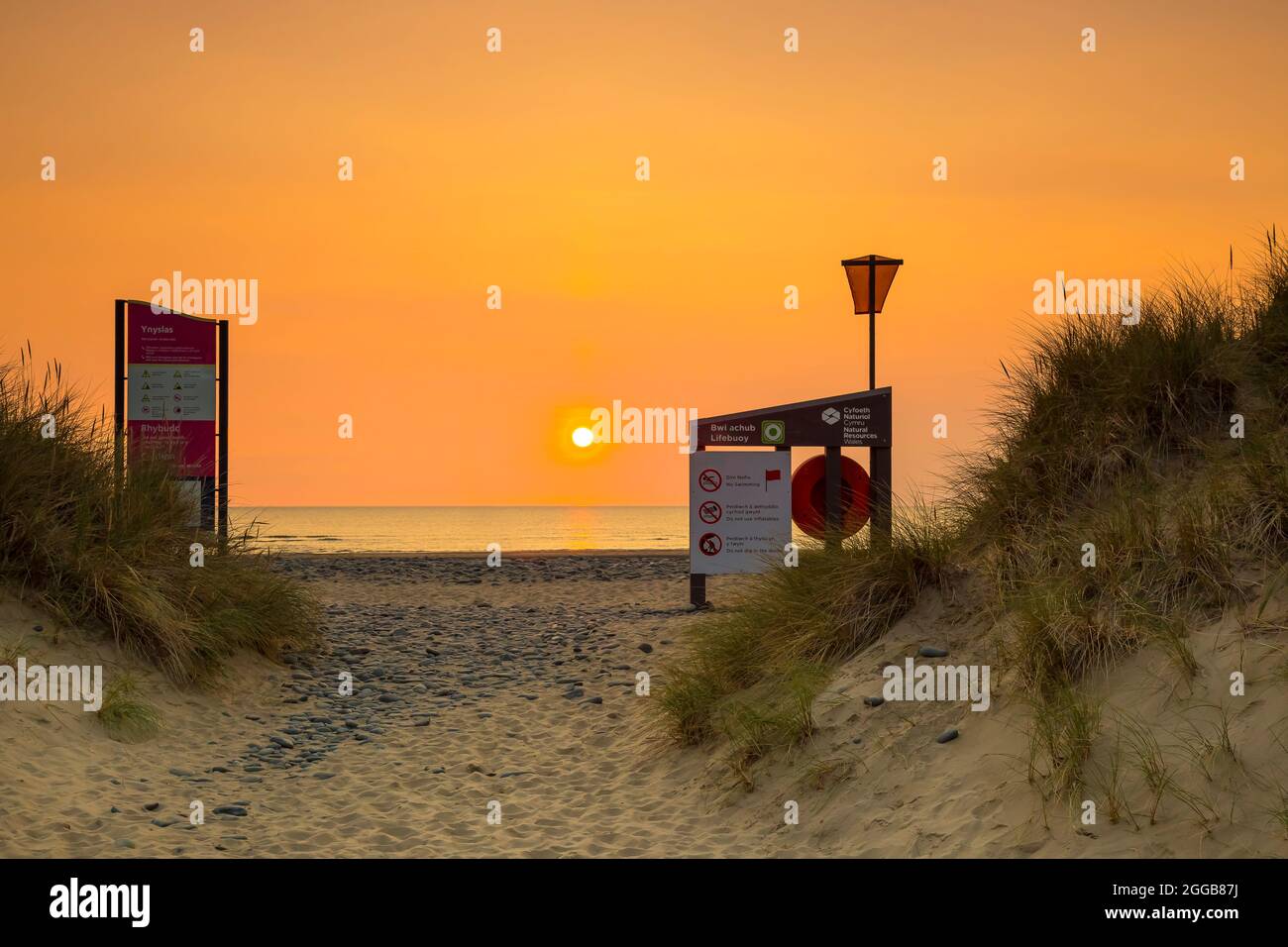 Blick zwischen diesen britischen Sanddünen, die auf eine untergehende Sonne über dem Meer blicken, mit Strandwarnschildern. Stockfoto