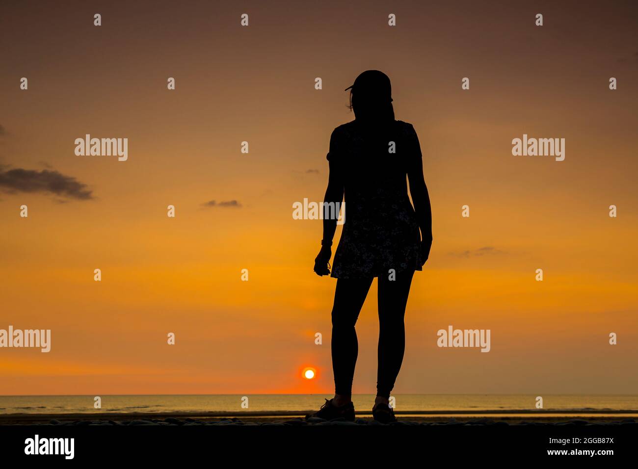 Rückansicht Silhouette einer isolierten jungen Frau, in Sportmütze, Blick auf das Meer bei Sonnenuntergang auf einem britischen Strand, Aufenthalt Sommerurlaub. Stockfoto