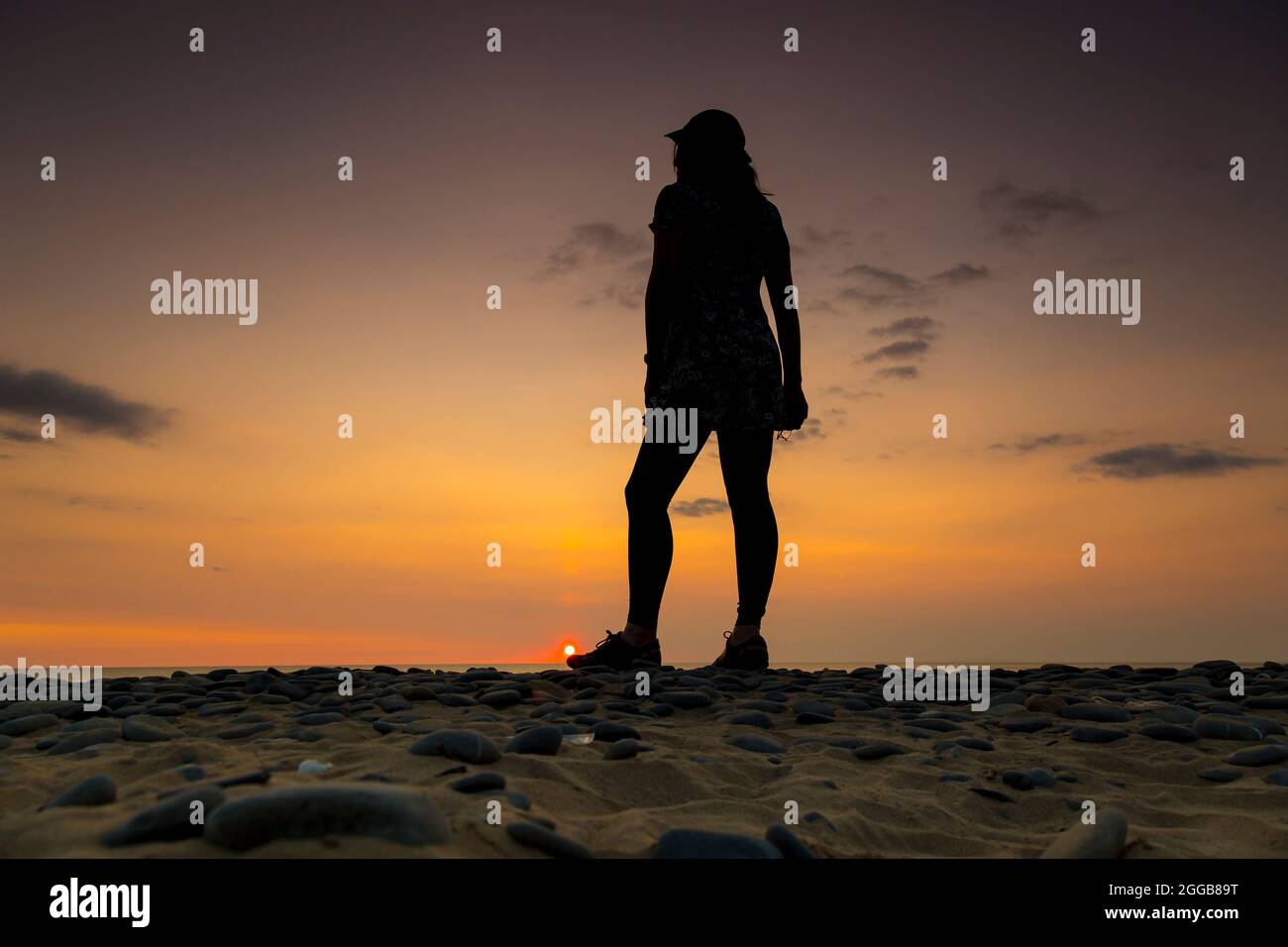 Rückansicht einer jungen Frau in Sportmütze mit Blick auf das Meer und den Sonnenuntergang in der Dämmerung auf einem britischen Strand, Aufenthalt im Sommerurlaub. Stockfoto