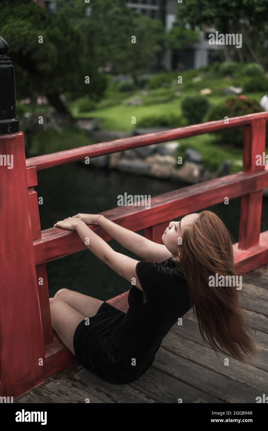 Porträt einer schönen jungen asiatischen Frau in lässigem schwarzen Kleid, die im Park auf der Brücke mit Handläufen sitzt. Schönes vietnamesisches Mädchen Stockfoto