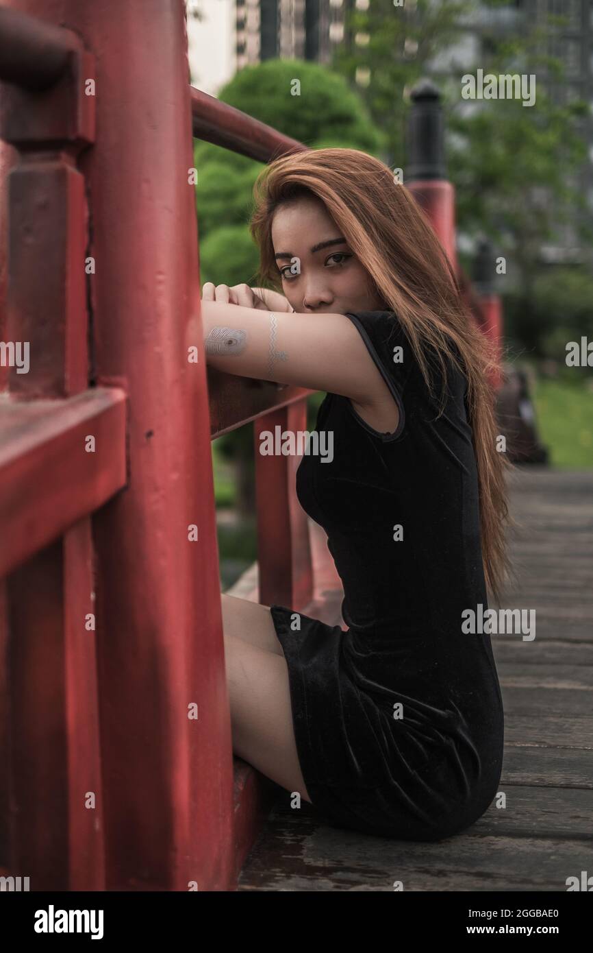 Porträt einer schönen jungen asiatischen Frau in lässigem schwarzen Kleid, die im Park sitzt und mit Kopierraum auf die Kamera schaut. Schönes vietnamesisches Mädchen Stockfoto