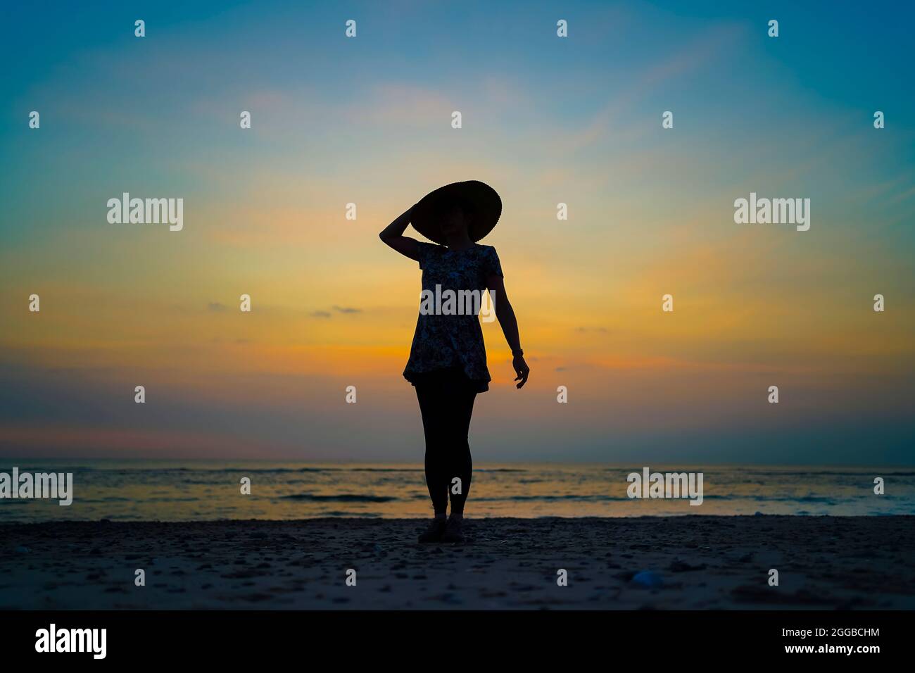 Silhouette einer jungen Frau bei Sonnenuntergang an einem britischen Strand, während der Sommerferien, einen Arm hoch, der sich an ihrem großen, schlampigen Sonnenhut festhält. Stockfoto
