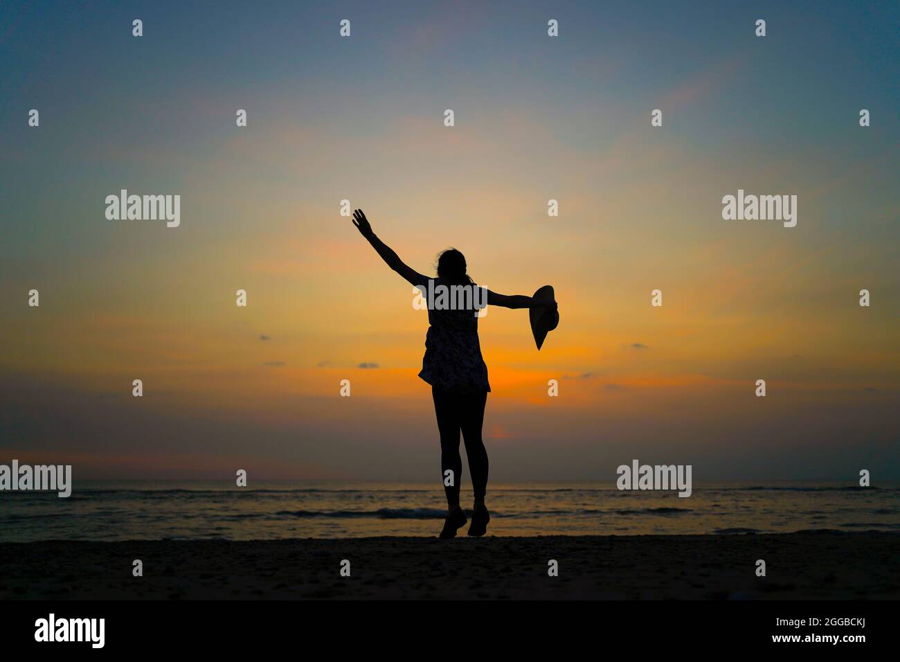 Rückansicht Silhouette einer jungen Frau, die bei Sonnenuntergang an einem britischen Strand tanzt, Sommerferien, ein Arm winkt in der Luft, ein Arm hält einen großen Sonnenhut. Stockfoto
