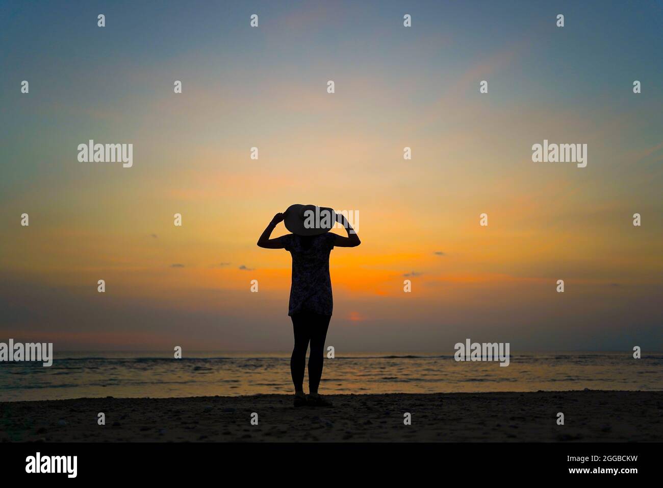 Rückansicht Silhouette einer jungen Frau mit Blick auf das Meer bei Sonnenuntergang an einem britischen Strand, Aufenthalt Sommerurlaub, hält sich an ihrem großen Floppy Sonnenhut. Stockfoto