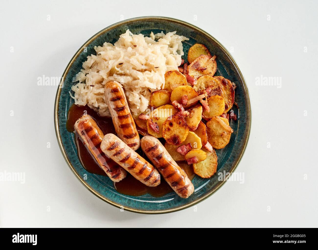 Würzige gegrillte oder gegrillte deutsche Würstchen mit gebratener Kartoffel- und Schinkenmischung und Weißkohl von oben auf einem Teller auf weißem Hintergrund Stockfoto