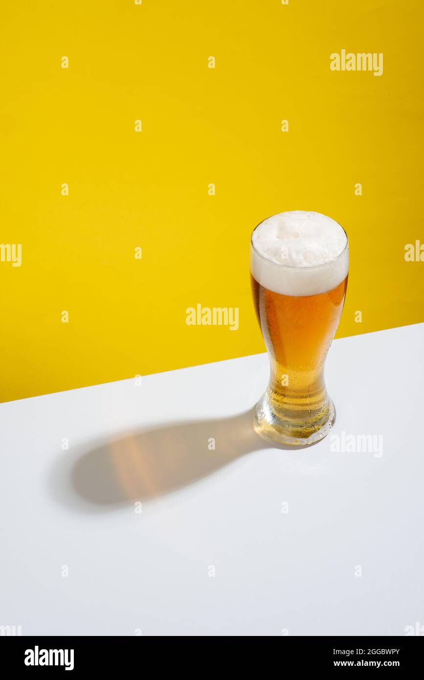 Kaltes Bier in einem Glas mit weißem Schaum auf einem weißen Tisch und gelbem Hintergrund serviert, keine Menschen Stockfoto