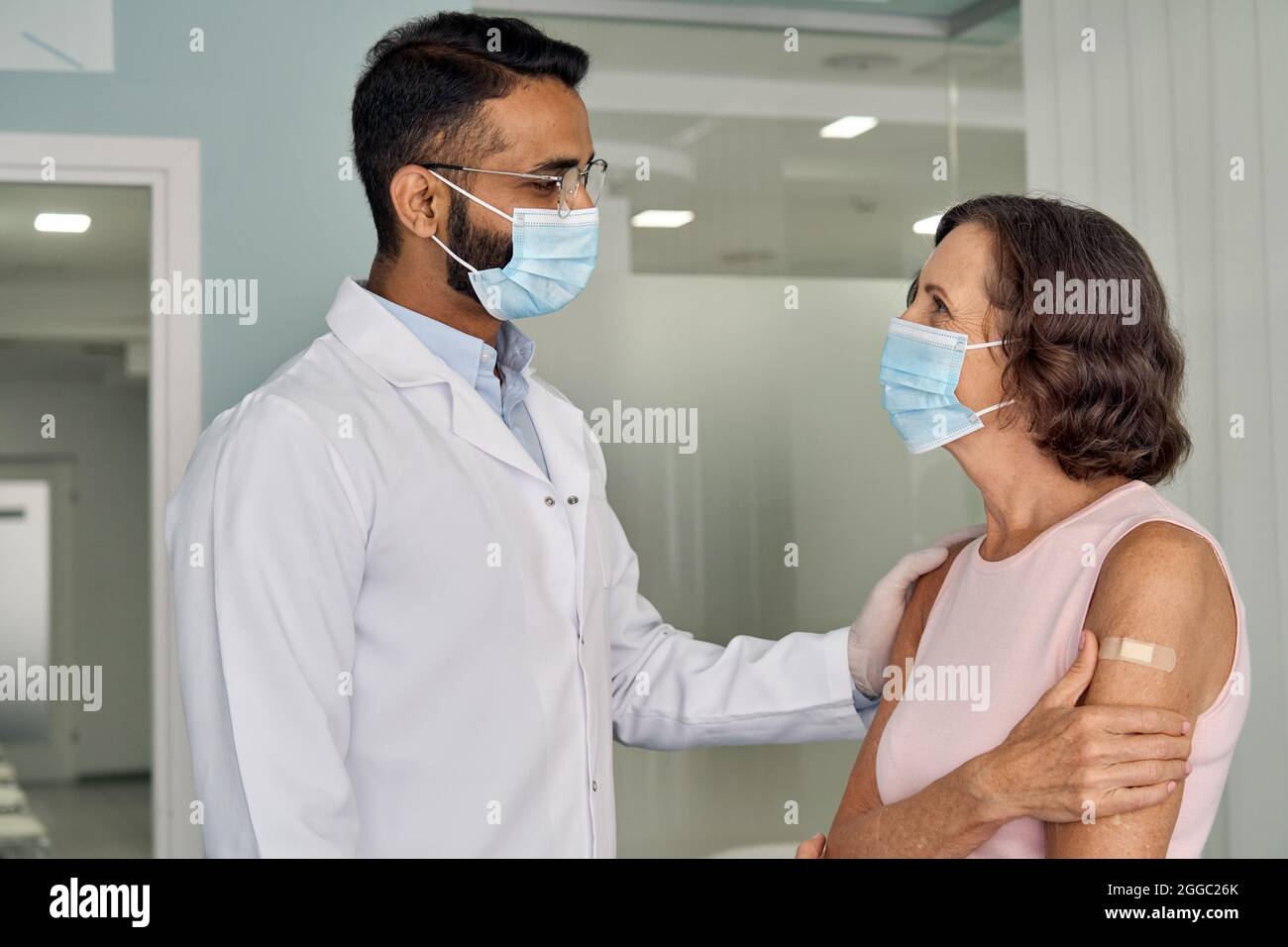 Fröhlicher indischer Arzt, der die ältere Patientin in Masken im Krankenhaus unterstützt. Stockfoto