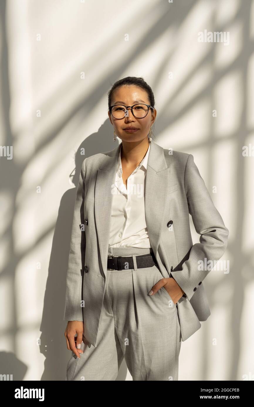 Junge, selbstbewusste Geschäftsfrau asiatischer Herkunft, die vor der Kamera gegen die weiße Wand steht Stockfoto