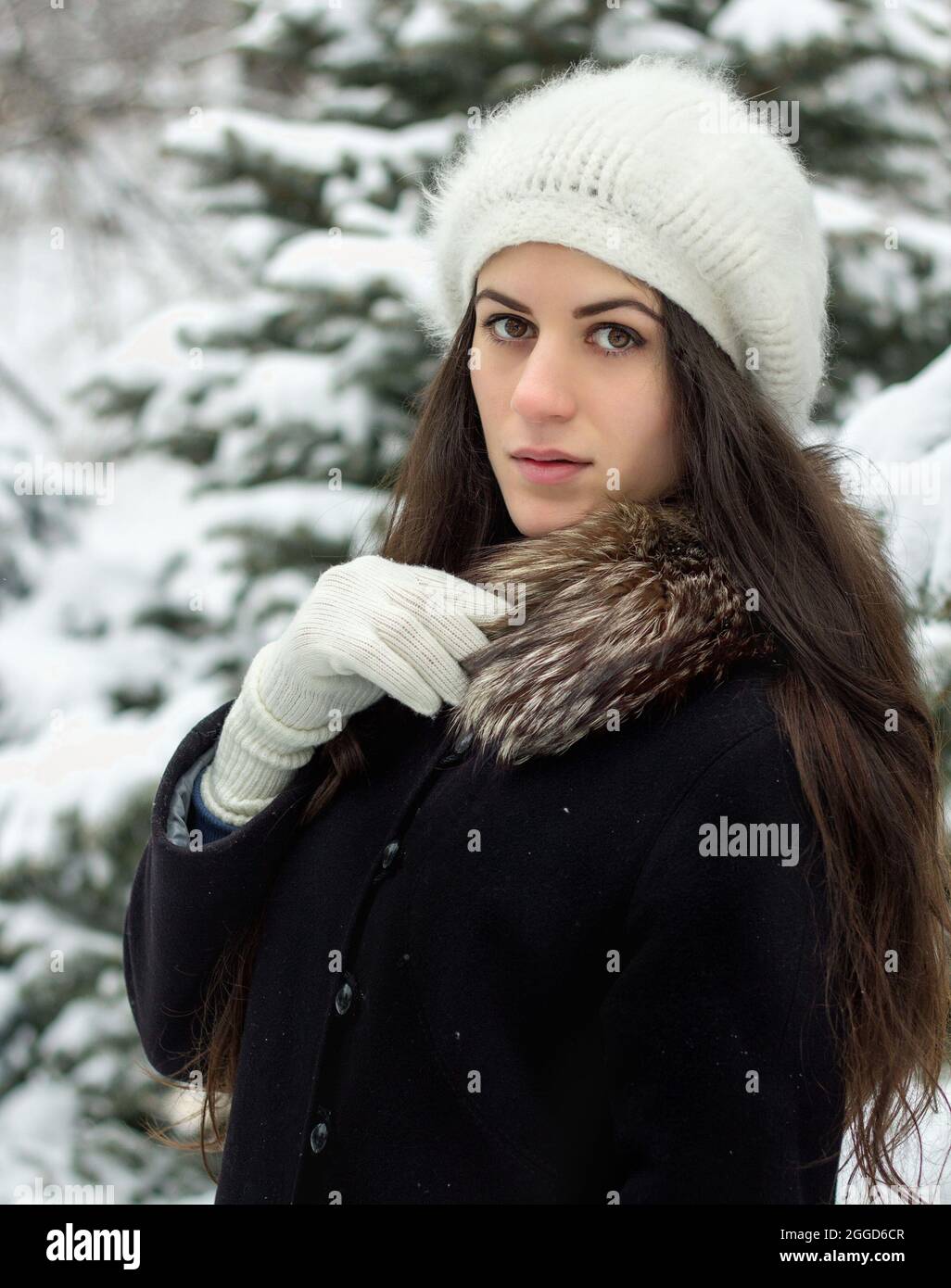 Fröhliche junge Frau, die draußen im Schnee in Strickmütze und weißen Handschuhen steht Stockfoto