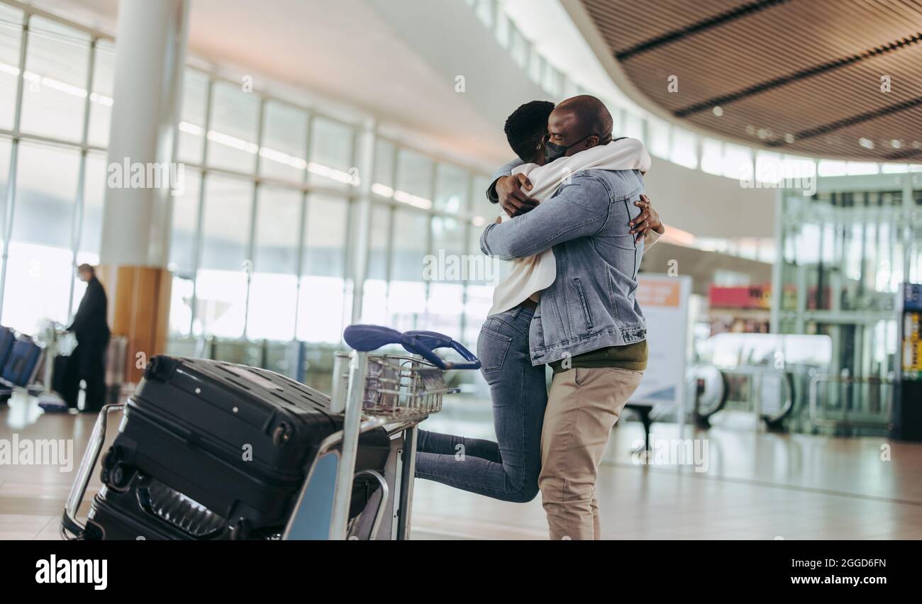 Mann mit Gesichtsmaske umarmt und hebt seine Frau am Flughafenterminal. Afrikanischer Mann trifft Frau, die am Flughafen ankommt. Stockfoto