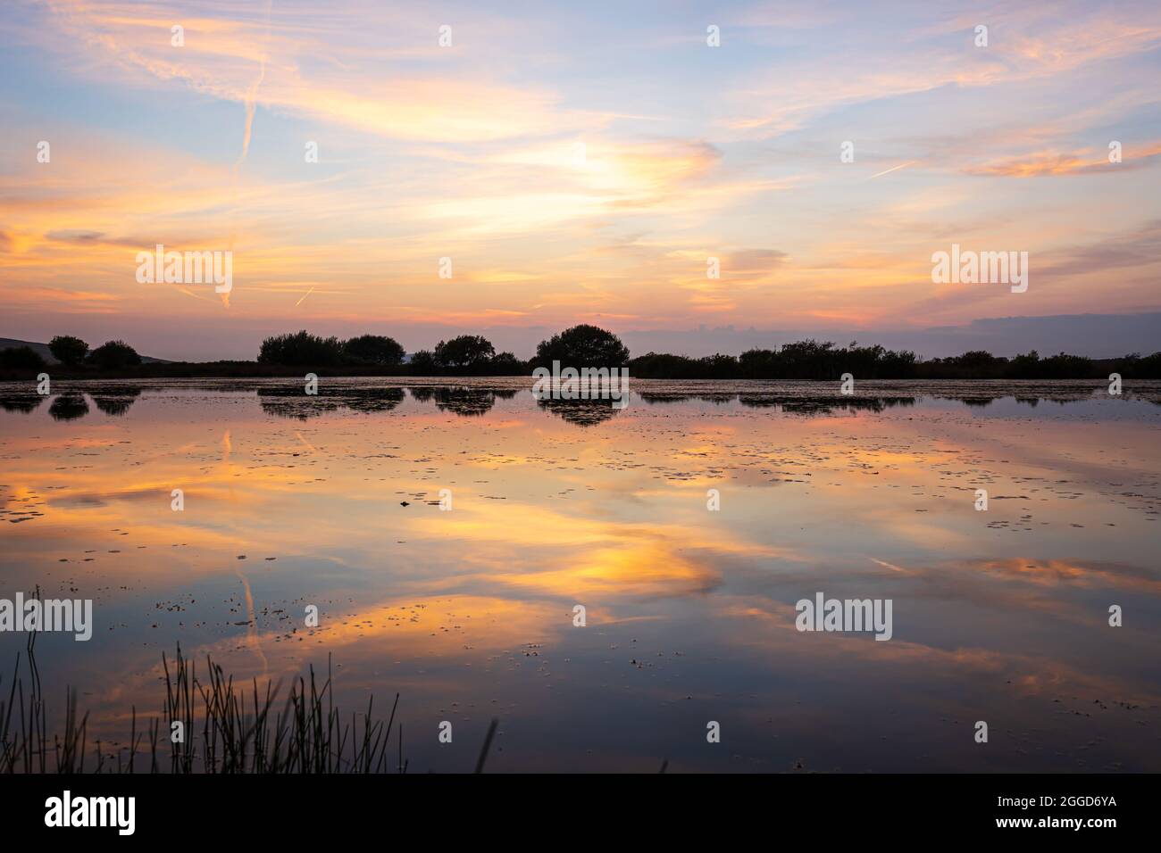 Schöner, heller Sonnenuntergang erleuchteter Himmel mit orangen Wolken mit Spiegelung im Wasser des Sees. Broad Pond, Gower Peninsula, Wales, Vereinigtes Königreich. Atemberaubende Sonnenuntergangslandschaft. Stockfoto