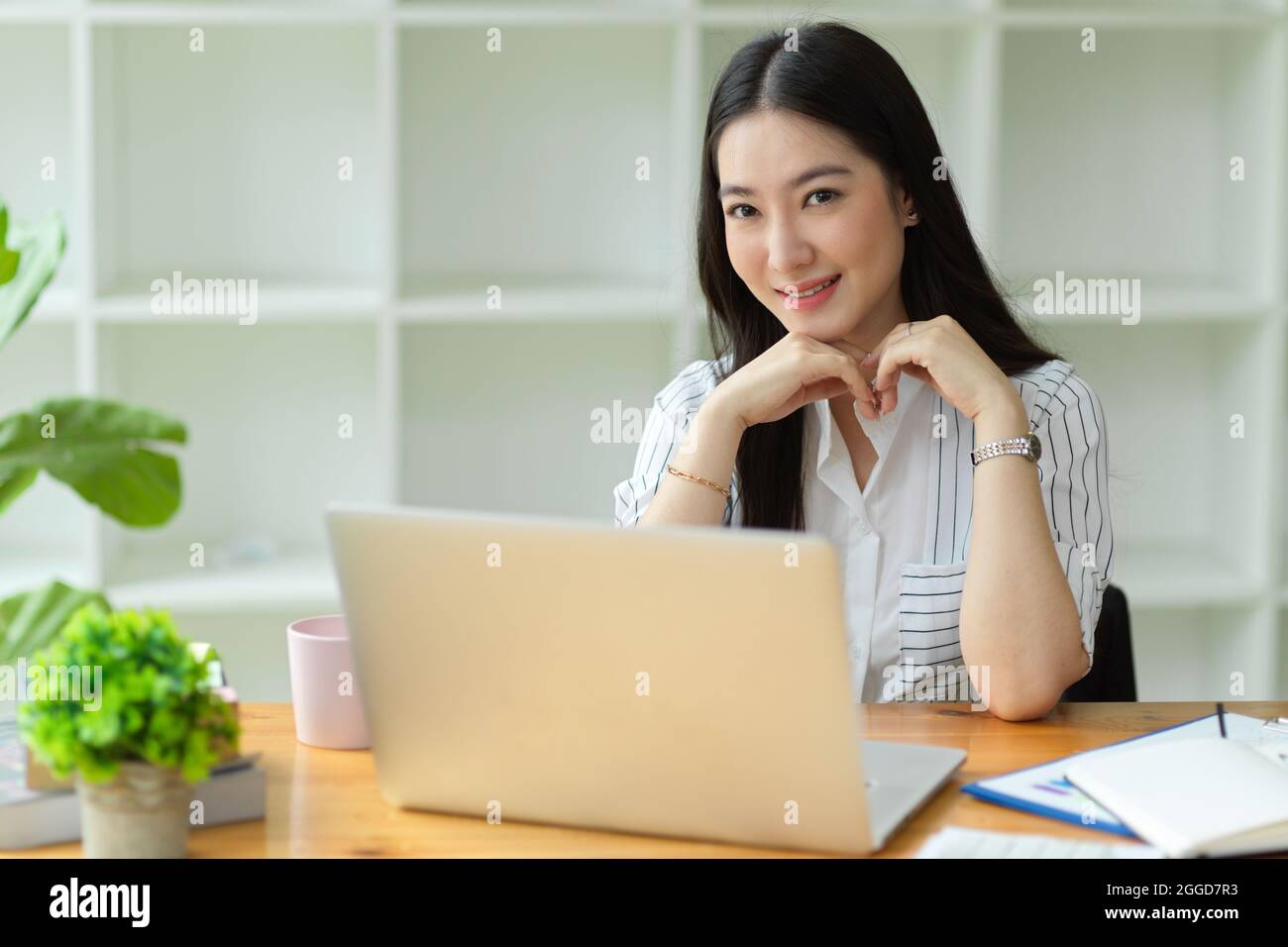Attraktive Geschäftsführerin lächelt und sitzt im Büro, Laptop, Bürobedarf, Zimmerpflanzen auf Holztisch Stockfoto