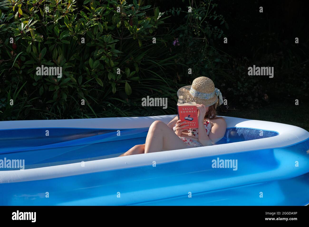 Eine junge Frau, die einen Sonnenhut trägt, liest ein Buch in einem Plastikplanschbecken in ihrem Garten Stockfoto