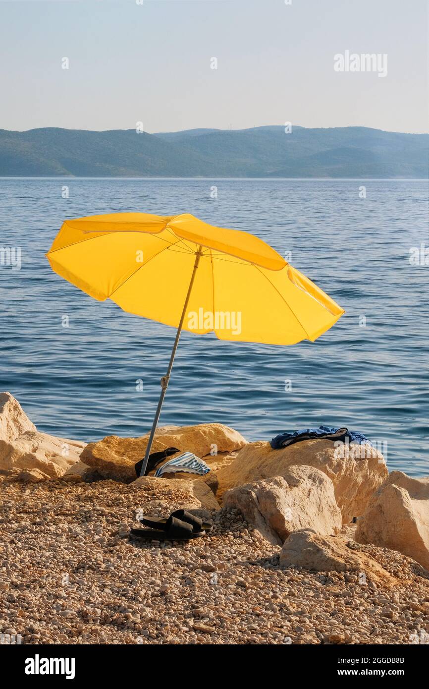 Gelber Sonnenschirm an der Sommerküste. Der Strand mit Sonnenschirm wartet auf Touristen am Sonnenuntergang. Happy Summer Resort Konzept. Vertikale Ansicht. Stockfoto