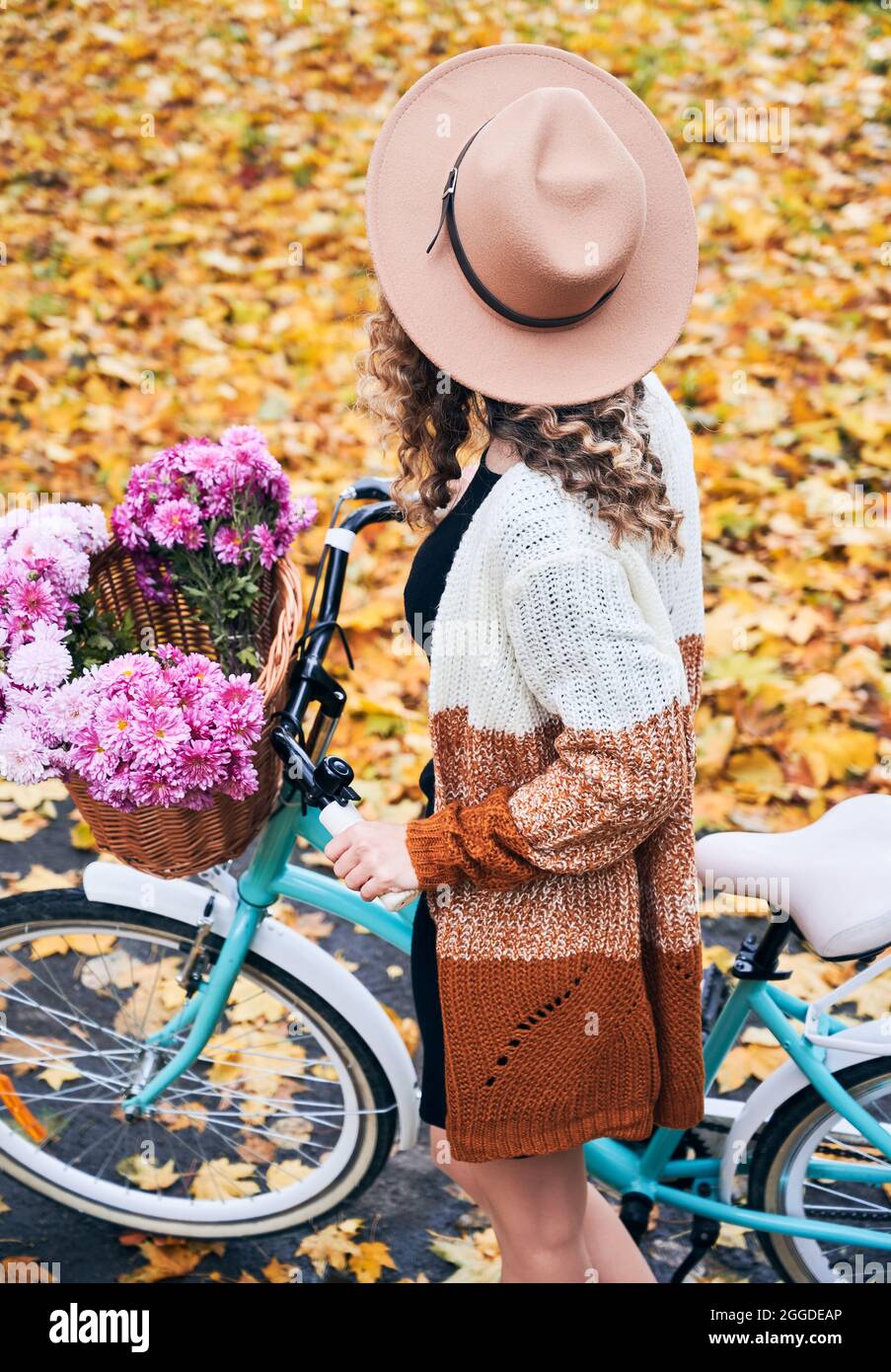 Oben Ansicht der jungen Frau mit lockigem Haar in beige Hut stehend mit blauen modernen Fahrrad und Strauß frischen rosa Blumen. Konzept des Spaziergangs auf dem Fahrrad im Herbstpark mit gelben Blättern. Stockfoto