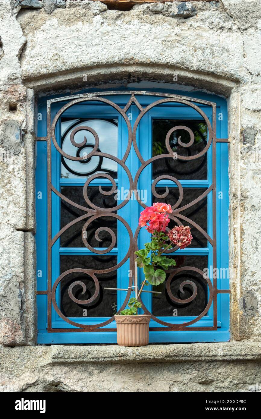 Dekoratives blaues Fenster mit kompliziertem Schmiedeeisen-Design und lebendiger Geranienpflanze in rustikaler Steinmauer, Auvergne Rhone Alpes, Frankreich Stockfoto