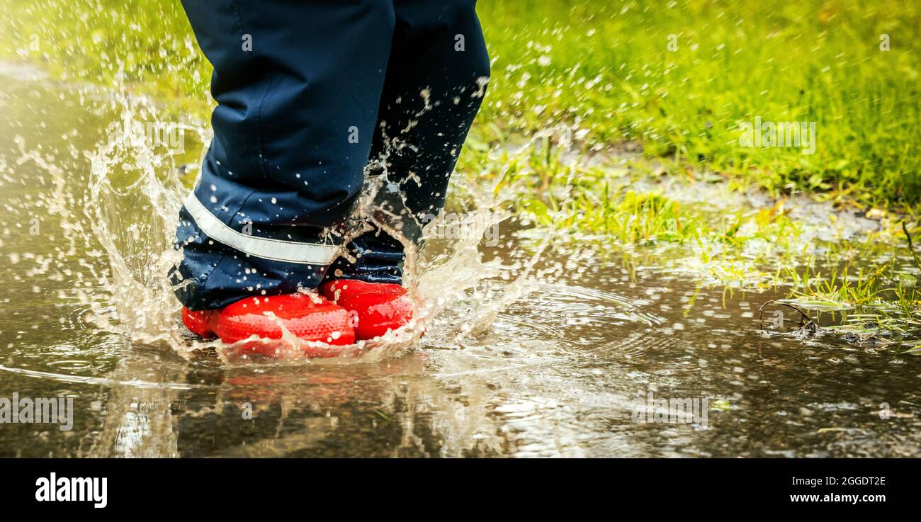Kind mit roten Gummistiefeln springen in Pfütze am regnerischen Herbsttag. Wasserdichte Kleidung. Copy Space Stockfoto