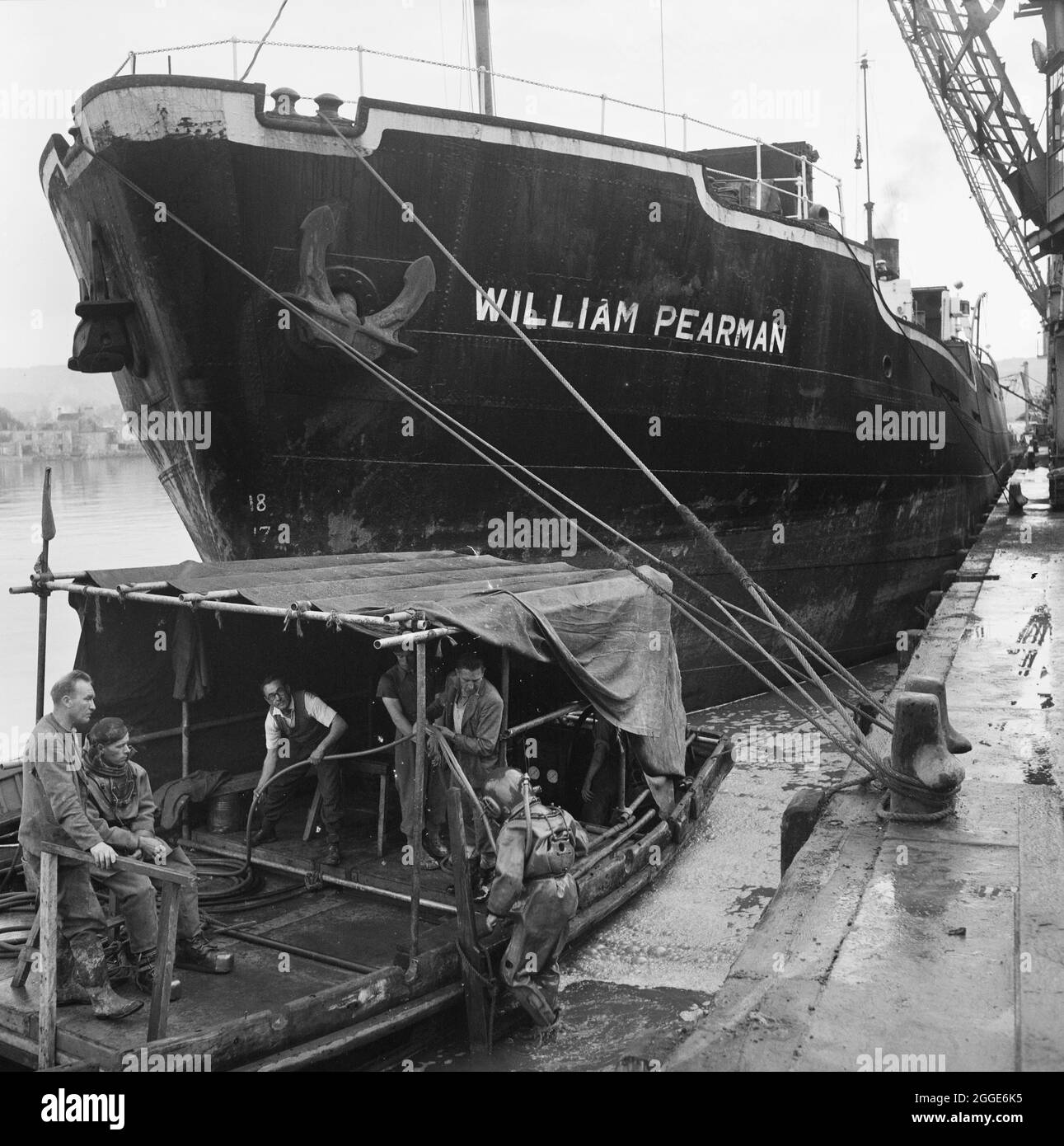 Ein Blick auf die SS William Pearman, die während des Baus am Coaling Wharf für das Plymouth 'B' Power Station festmachte, zeigt Arbeiter an Bord eines kleineren Schiffes im Vordergrund mit einem Taucher, der ins Wasser eindringt. Dieses Bild wurde als Teil des Breaking New Ground Project in Zusammenarbeit mit dem John Laing Charitable Trust in den Jahren 2019-20 katalogisiert. Stockfoto