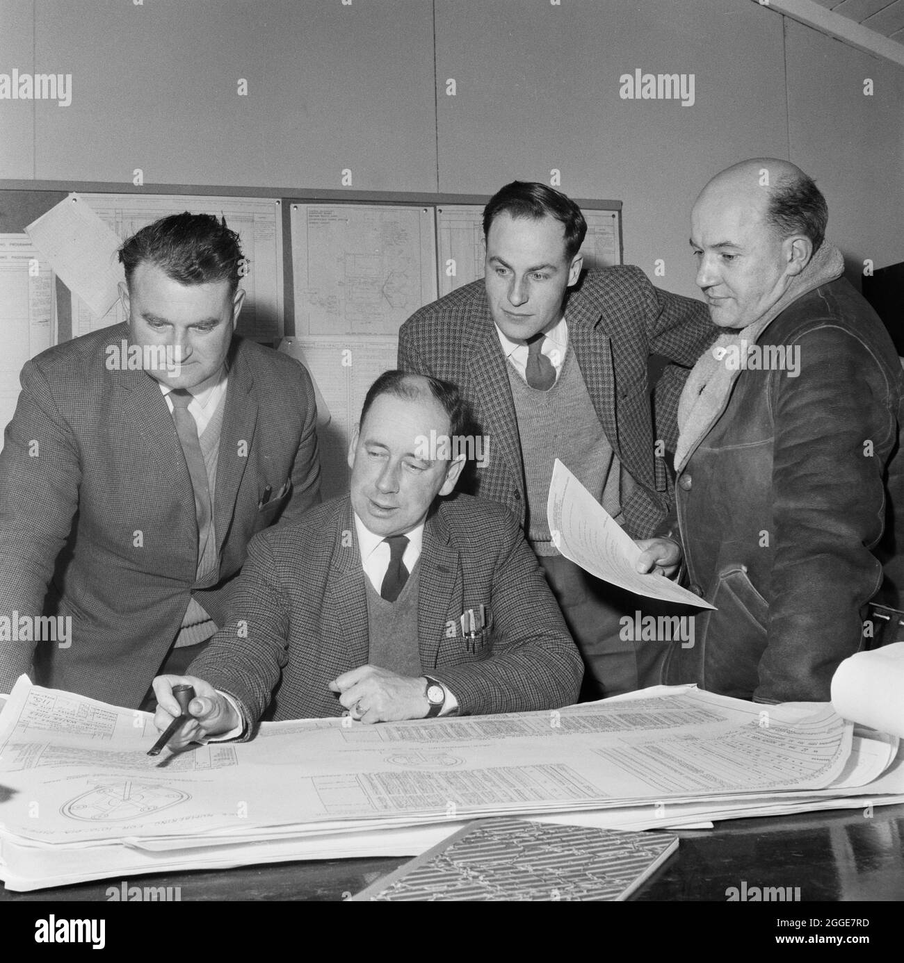 Vier Laing-Arbeiter, D. S. Smith, J. C. Proud, M. J. Taylor und J. P. Lapping, schauten sich Pläne für die Belastung von Kühlwasserleitungen während des Baus des Kraftwerks Eggborough an. Eine ähnliche Version dieses Bildes wurde in der Mai 1963 Ausgabe des monatlichen Laing-Newsletters Team Spirit veröffentlicht. Die Beschriftung lautet „D. S. Smith (sitzend), Agent, bespricht die Belastung von Kühlwasserrohren mit J. C. Proud, Construction Agent (links), M. J. Taylor, Production Controller, und J. P. Lapping, Senior Engineer (ganz rechts).“ Stockfoto