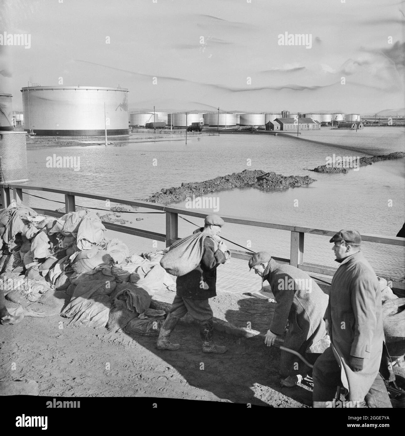 Überschwemmungen auf der Baustelle der Coryton Ölraffinerie, die Hochwasser im Tanklagerbereich und Arbeiter, die Sandsäcke transportieren, im Vordergrund zeigen. In der Nacht vom 31. Januar bis zum Morgen des 1. Februar 1953 ereigneten sich schwere Überschwemmungen in Teilen Großbritanniens, Schottlands, Belgiens und der Niederlande. Die Naturkatastrophe wird oft als die Nordseeflut von 1953 bezeichnet. An der Ostküste Großbritanniens kam es zu schweren Überschwemmungen. Der Standort der Coryton Ölraffinerie überflutete, als an 14 Stellen die Flussmauer zum Schutz des Standorts durchbrochen wurde. Ein Artikel, der die Katastrophe und die Unterbesatzungen beschreibt Stockfoto